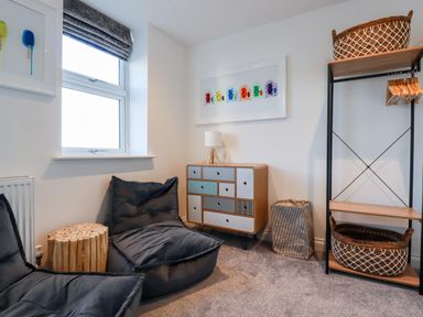 A cozy room with two gray cushioned chairs a small wooden side table a patterned drawer unit lamp shelving with baskets and hangers and colorful artwork on white walls at Chy Fistral in Newquay