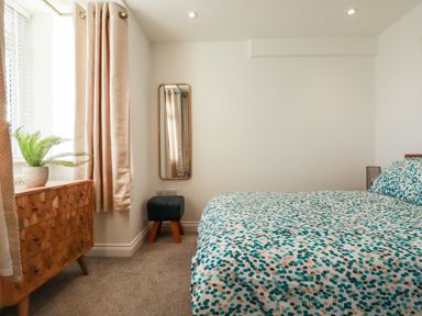A bedroom with a bed with patterned bedding a wooden dresser with a plant a window with curtains a wall mirror and a small upholstered stool at Chy Fistral in Newquay