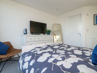 A bedroom with a bed a dresser with a television a woven chair and a full length mirror at Chy Fistral in Newquay