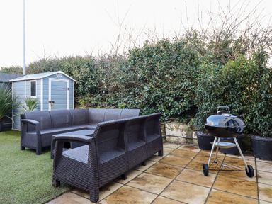 A patio with wicker outdoor furniture a charcoal grill a small shed and bushes at Chy Fistral in Newquay