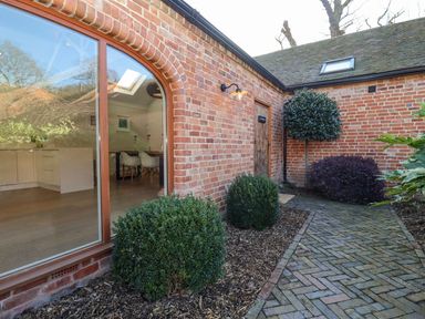 An outdoor brick walkway next to a building with large windows showing a kitchen and dining area at The Piggery in Ledbury near Welland