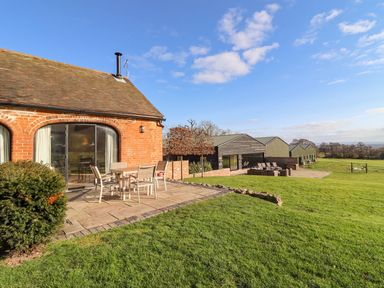A brick building with glass doors and patio furniture on a stone patio next to a lawn with additional outbuildings at The Piggery in Ledbury near Welland