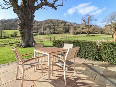 An outdoor patio with a square table and four chairs overlooking a fenced grassy field with large trees at The Piggery in Ledbury near Welland