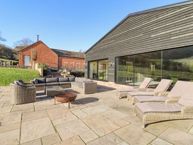 An outdoor patio with cushioned seating a fire pit and lounge chairs next to a black wooden building at The Piggery in Ledbury near Welland