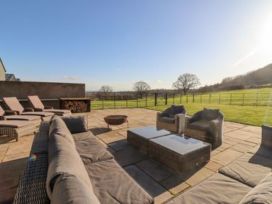 An outdoor patio with cushioned wicker sofas chairs and a fire pit overlooking a grassy fenced field at The Piggery in Ledbury near Welland