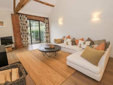 A living room with a white sectional sofa, cushions, round black coffee table, TV on a stand, and sliding glass door at The Piggery in Ledbury near Welland