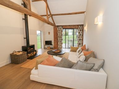 A living room with a white sofa and cushions a wood stove and basket with logs a round coffee table and glass doors at The Piggery in Ledbury near Welland