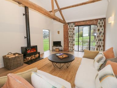 A living room with a wood burning stove a round coffee table a white sofa with cushions and sliding glass doors at The Piggery in Ledbury near Welland