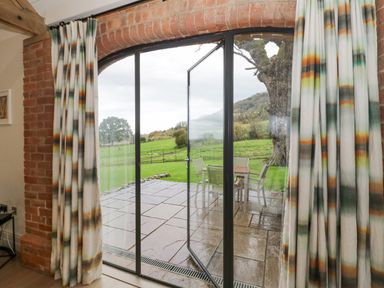 A patio door with patterned curtains opening to a stone patio with outdoor furniture and a grassy field beyond at The Piggery in Ledbury near Welland