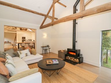 A living room with a white sofa and cushions around a round black coffee table a wood stove a basket of firewood and visible wooden beams at The Piggery in Ledbury near Welland