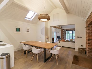 A dining area with wooden table and white chairs next to a living room with a sofa and wood burning stove at The Piggery in Ledbury near Welland
