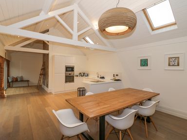 A dining area with a wooden table and white chairs next to a kitchen with white cabinets and appliances at The Piggery in Ledbury near Welland