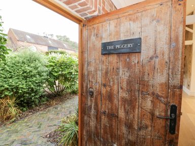 A wooden door with a sign reading The Piggery opening to a garden with bushes and a brick pathway at The Piggery in Ledbury near Welland