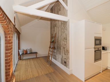 A kitchen area with white cabinets and built-in oven next to a small seating area with a bench and pillows at The Piggery in Ledbury near Welland