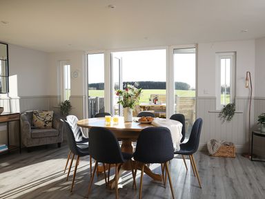 A dining area with a round wooden table and six chairs near large windows showing a field at Ash Cottage in Lucker near Warenford