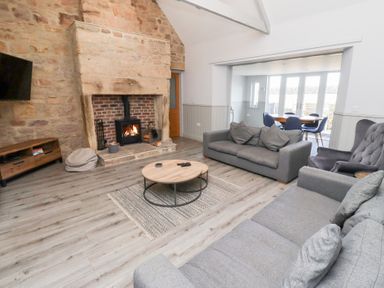 A living room with a large stone fireplace and wood burning stove several grey sofas a round wooden coffee table and a dining area with blue chairs at Ash Cottage in Lucker near Warenford