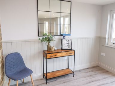A corner of a room with a blue chair a wooden console table with decor and a square grid mirror on the wall at Ash Cottage in Lucker near Warenford