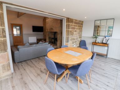 A dining area with a wooden round table and six blue chairs next to a living room with a grey sofa and stone fireplace at Ash Cottage in Lucker near Warenford
