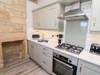 A kitchen with gray cabinets a built in oven a gas stove a toaster a microwave and stone wall at Ash Cottage in Lucker near Warenford