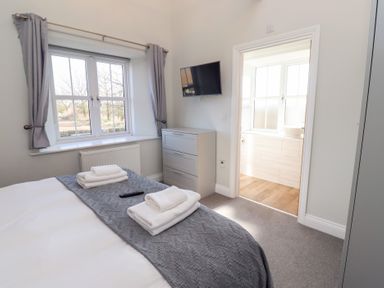 A bedroom with a bed, folded towels on the bed, a window with curtains, a chest of drawers, a wall-mounted TV and an open door leading to a bathroom at Ash Cottage in Lucker near Warenford