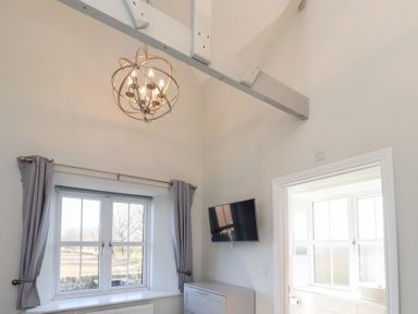 A room with a window with curtains a hanging light fixture a wall-mounted television and a dresser at Ash Cottage in Lucker near Warenford