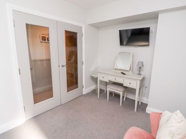 A room with double glass panelled doors a white dressing table with a mirror stool and lamp a wall mounted television and a pink armchair with a cushion at Ash Cottage in Lucker near Warenford