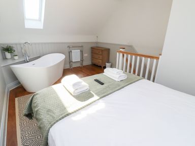 A bedroom with a bed having folded towels and a remote a bathtub near a skylight window a wooden chest of drawers and a wooden stair railing at Ash Cottage in Lucker near Warenford
