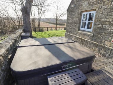 An outdoor hot tub with a closed cover on a wooden deck next to a stone building with a window at Ash Cottage Lucker near Warenford