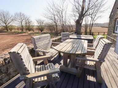 An outdoor seating area with wooden benches and chairs around a wooden table on a deck at Ash Cottage in Lucker near Warenford