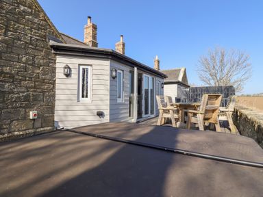An outdoor patio with wooden chairs and a table next to a building with stone and light grey siding at Ash Cottage in Lucker near Warenford