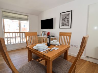 A dining area with a wooden table set for two with wine glasses and a bottle of red wine a wall-mounted television and framed artwork at Skylark in Southwold
