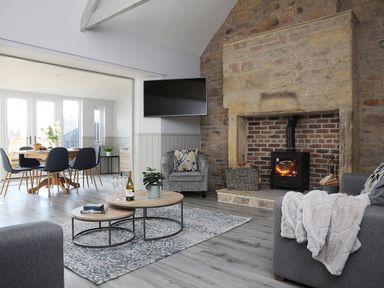 A living room with a stone fireplace, a wood burning stove, a wall-mounted TV, grey sofas, and a dining area with a round wooden table and chairs at Elm Cottage in Lucker near Warenford