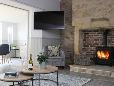 A living room with a stone fireplace and wood burner a round coffee table with wine glasses a wine bottle and a plant an armchair a wall mounted television at Elm Cottage in Lucker near Warenford