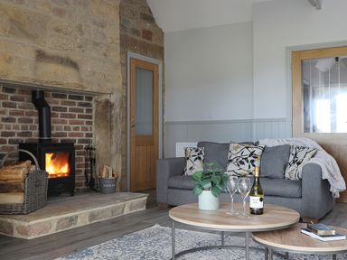 A living room with a stone fireplace a wood burning stove a grey sofa with patterned cushions and a wooden coffee table with a plant wine bottle and glasses at Elm Cottage in Lucker near Warenford