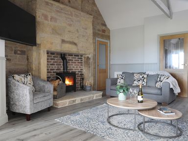 A living room with a stone fireplace a gray sofa and armchair a wooden coffee table and a television mounted on the wall at Elm Cottage in Lucker near Warenford