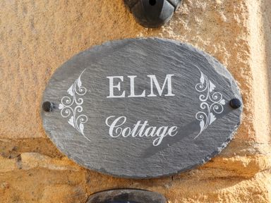 A grey slate oval house sign with the words Elm Cottage mounted on a stone wall at Elm Cottage in Lucker near Warenford