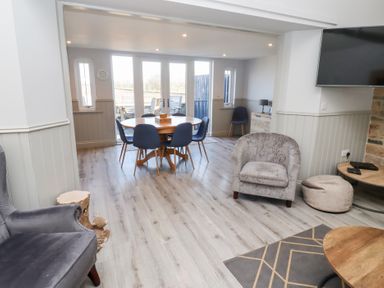 A dining area with round wooden table and six chairs and a sitting area with armchairs and a wall-mounted TV at Elm Cottage in Lucker near Warenford