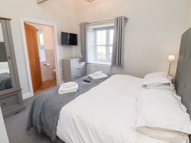 A bedroom with a bed with white linens a grey blanket folded towels a chest of drawers a wall mounted tv and a window with curtains at Elm Cottage in Lucker near Warenford