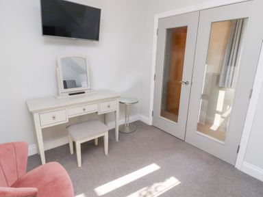A room with a white dressing table with mirror a small stool a wall-mounted tv a pink chair and double doors with glass panels at Elm Cottage in Lucker near Warenford