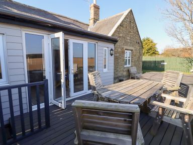 An outdoor deck area with wooden table and chairs outside a house with glass doors and stone walls at Elm Cottage in Lucker near Warenford