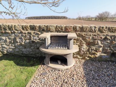 A stone outdoor barbecue grill against a stone wall with a field and trees in the background at Elm Cottage in Lucker near Warenford