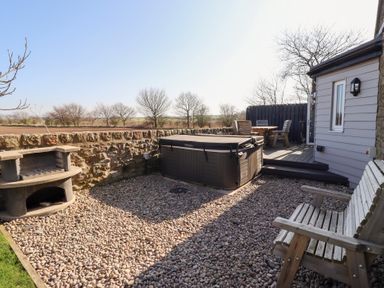 An outdoor patio area with a hot tub a wooden bench a stone barbecue and a wooden table with chairs at Elm Cottage in Lucker near Warenford