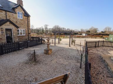 A gravel courtyard with a bench a fountain a wooden arch and a stone house at Elm Cottage in Lucker near Warenford