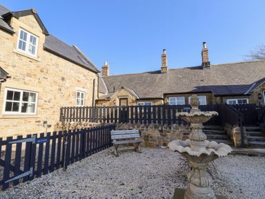 An outdoor courtyard with a stone fountain a wooden bench gravel ground and stone cottages at Elm Cottage in Lucker near Warenford