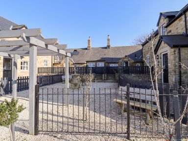 An outdoor area with stone buildings a metal gate benches a wooden pergola and gravel ground at Elm Cottage in Lucker near Warenford