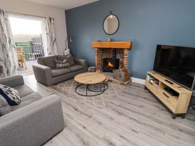 A living room with two grey sofas a round wooden coffee table a fireplace with a wood burning stove a wall mirror and a large TV on a wooden stand at Oak Cottage in Lucker near Warenford