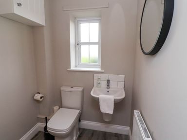 A small bathroom with a toilet sink a round mirror and a window at Oak Cottage in Lucker near Warenford