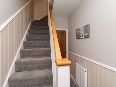 A carpeted staircase with a wooden handrail next to a wall with three floral paintings and a radiator at Oak Cottage in Lucker near Warenford