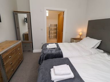 A bedroom with two single beds with white bedding and gray blankets a wooden dresser a cabinet with a mirror a bedside table with a lamp and an open door showing a bathroom Oak Cottage Lucker near Warenford