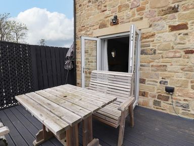 An outdoor patio with a wooden table and bench adjacent to an open door in a stone wall at Oak Cottage in Lucker near Warenford
